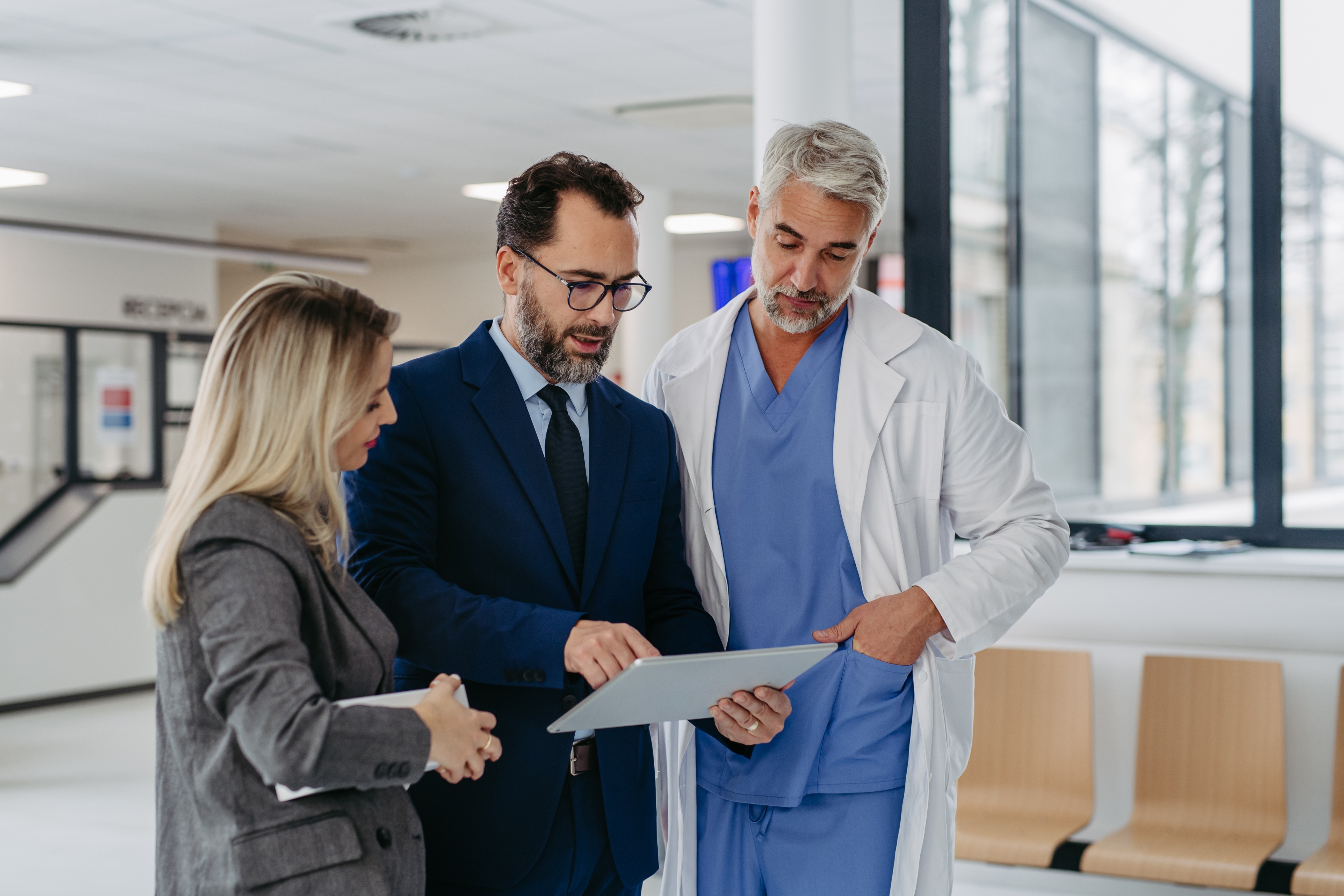 A man holding a tablet in a hospital setting with a doctor and a woman looking at the tablet screen