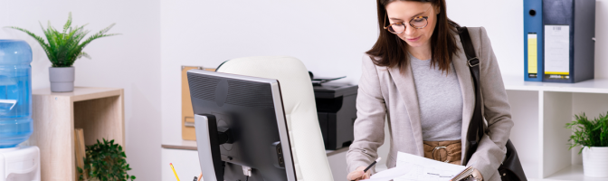 businesswoman with handbag on shoulder looking through papers while leaning over desk in office