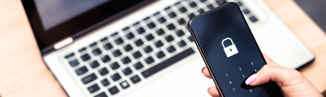Hand holding a phone hovering over laptop computer with a password lock on the screen. 