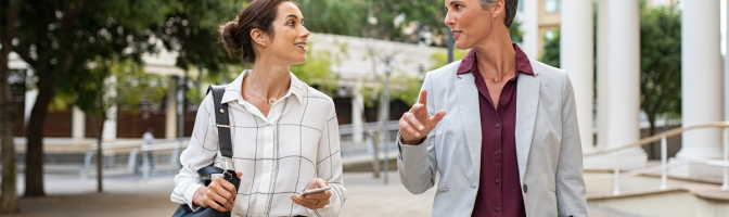 Two business women in conversation walking on city street.