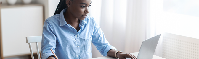 Woman Working On Laptop At Home Office And Taking Notes, Sitting At Desk Near Window, Looking At Screen, Writing Down Information From Computer