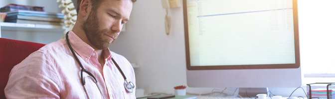 Side view of a Caucasian male doctor checking medical paper in clinic against his computer in background