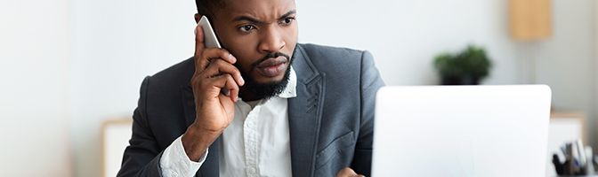 Business owner sitting in front of a laptop while holding a phone to his ear.