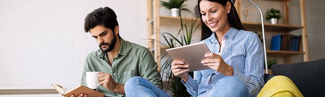 A woman reads an eBook while a man reads a book as he holds a cup in one hand.