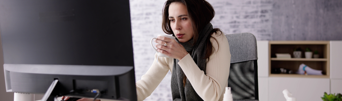 Sick female employee sitting at desk drinking tea while working.