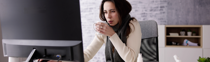 Sick female employee sitting at desk drinking tea while working. 