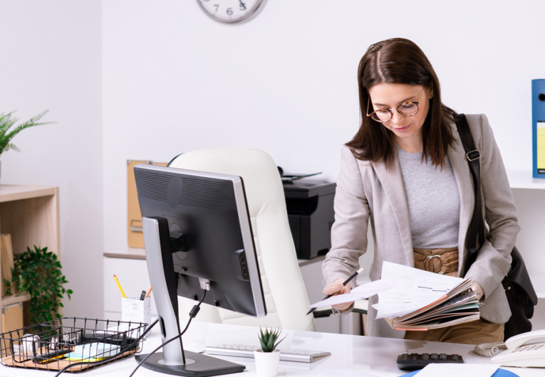 businesswoman with handbag on shoulder looking through papers while leaning over desk in office