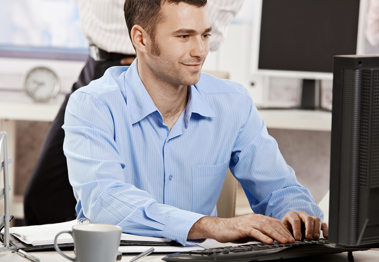 Casual businessman working in office, sitting at desk, typing on keyboard, looking at computer screen.