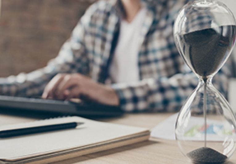 Blurred image of a person working at a computer with an hourglass in the forefront.