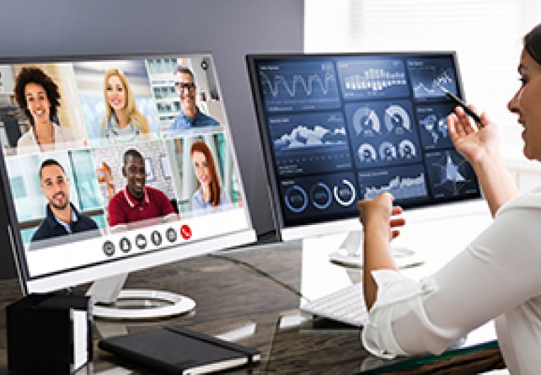 Business woman participating in a virtual conference, sitting in front of two computer screens that show attendees.