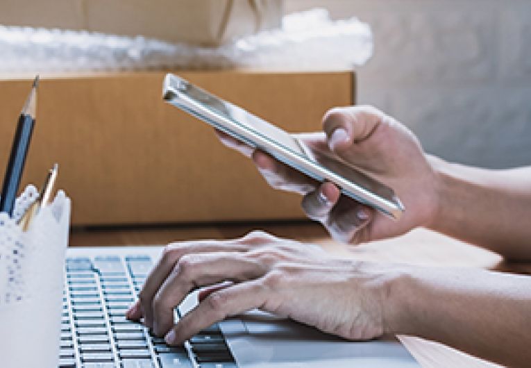 Business owner sitting at a desk in front of a laptop while looking at a mobile phone.