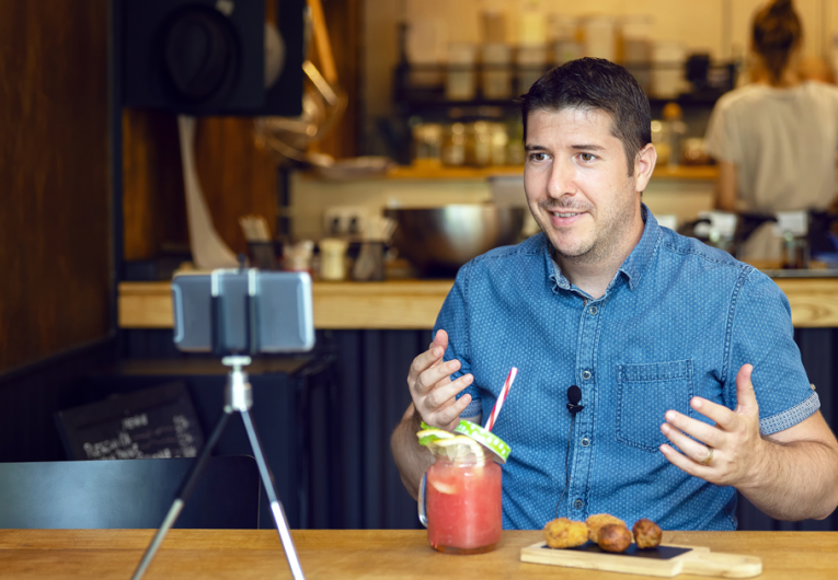 Social media influencer or food blogger creating content inside small restaurant. Man sharing online food review using smartphone on tripod. 
