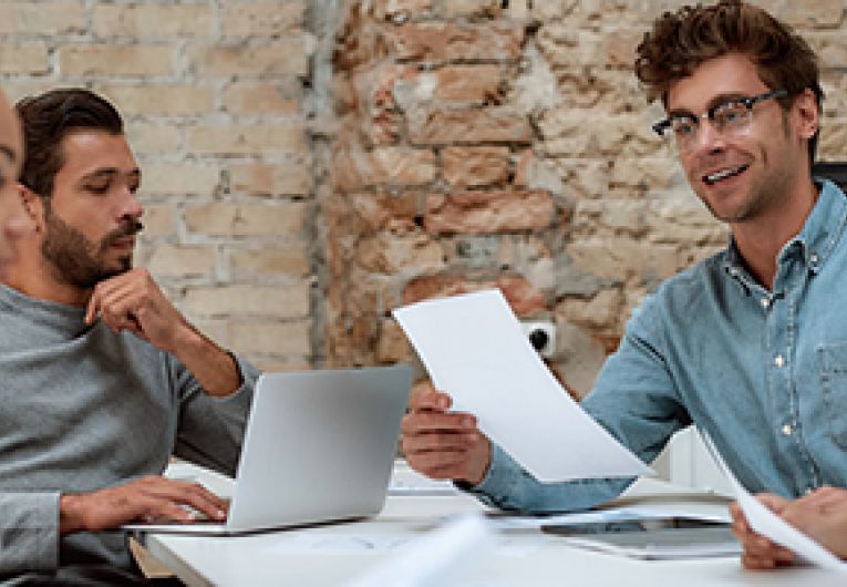 A group of six business professionals have a discussion around a meeting table.