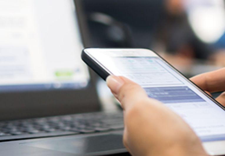Close up of a person typing into a smart phone in front of a laptop.