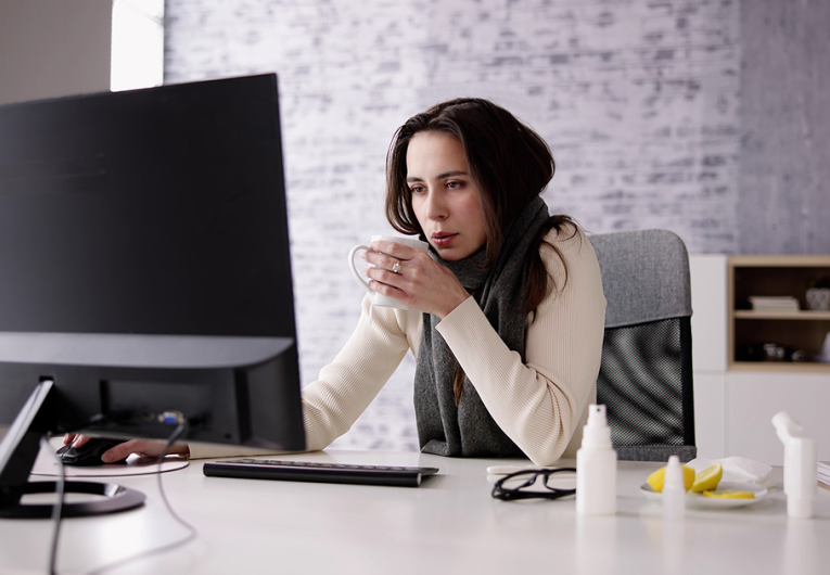 Sick female employee sitting at desk drinking tea while working. 