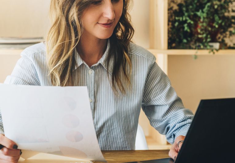 younger woman with light hair references document and laptop at desk