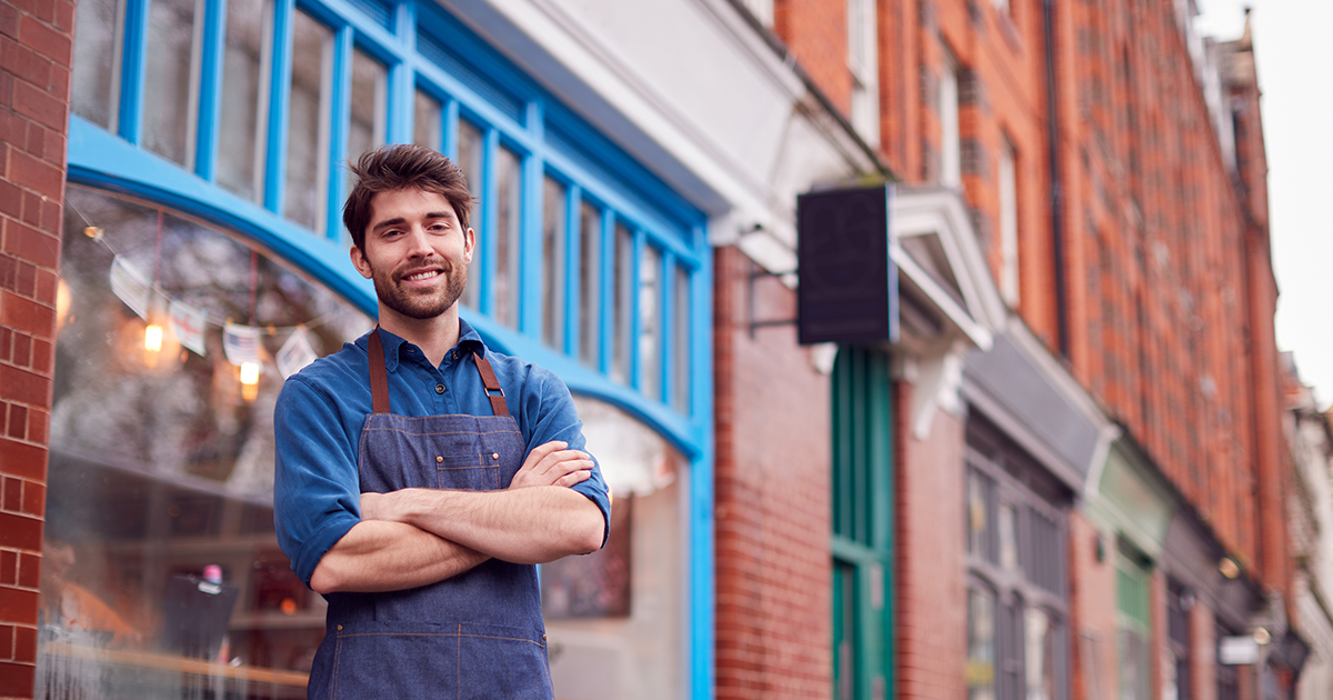 Portrait Of Male Small Business Owner Wearing Apron Standing Outside Shop On Local High Street