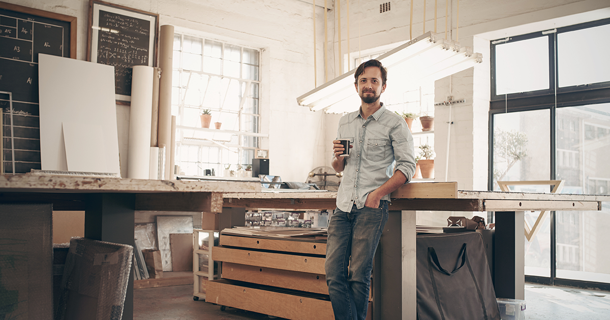 young male designer standing comfortably in his naturally lit workshop, holding his morning cup of coffee and smiling at the camera