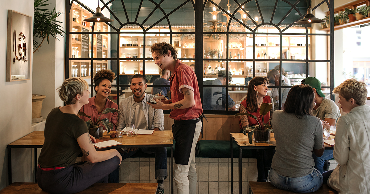 Smiling young waiter taking orders from a diverse group of customers sitting together at a restaurant table