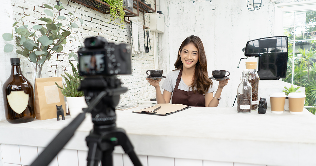 young female barista in apron smiling with recording video camera filming at coffee shop counter.