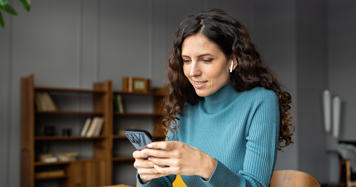 Smiling woman sitting at office desk looking at phone with earbuds in ears listening to podcast. 