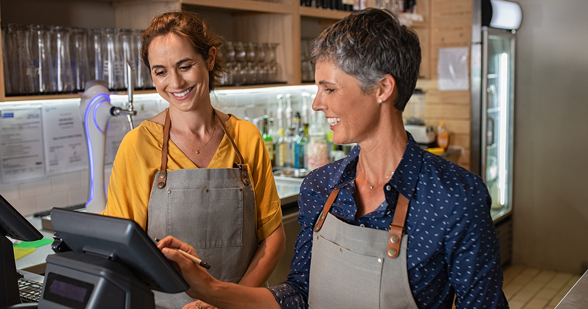 Two happy coffeehouse waitresses in apron smiling and working on cash register.